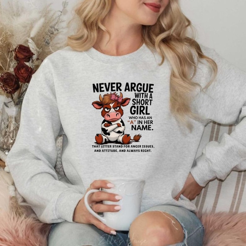 May include: A light gray sweatshirt with the text "NEVER ARGUE WITH A SHORT GIRL WHO HAS AN 'A' IN HER NAME." A cartoon cow with a pink bow is featured in the center. The text below reads "THAT LETTER STAND FOR ANGER ISSUES, AND ATTITUDE, AND ALWAYS RIGHT."