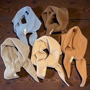 Puede incluir: Cinco bufandas de punto suave en varios colores neutros: azul claro, marrón, beige y marrón claro. Cada bufanda tiene una etiqueta y está colocada sobre una superficie de madera oscura. Las bufandas están diseñadas para llevarse alrededor del cuello.