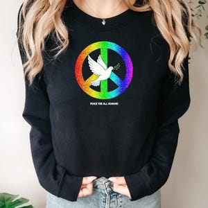 Könnte beinhalten: Schwarzes Sweatshirt mit einem Regenbogen-Friedenssymbol und einer weißen Taube mit einem Olivenzweig. Der Text "Peace for all humans" ist darunter aufgedruckt. Ein lässiges, bequemes Oberteil.