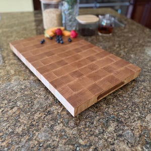 Puede incluir: Una tabla de cortar rectangular de madera de grano final con un patr&oacute;n de cuadr&iacute;cula de bloques de madera de color marr&oacute;n claro y medio. La tabla tiene un perfil grueso con una superficie lisa y una ranura de agarre lateral para facilitar su uso.
