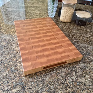Puede incluir: Una tabla de cortar rectangular de madera de testa con un patr&oacute;n de cuadros de color marr&oacute;n claro. La tabla tiene una superficie lisa y un asa recortada horizontal en el lateral, mostrando la veta natural de la madera en cuadr&iacute;cula.