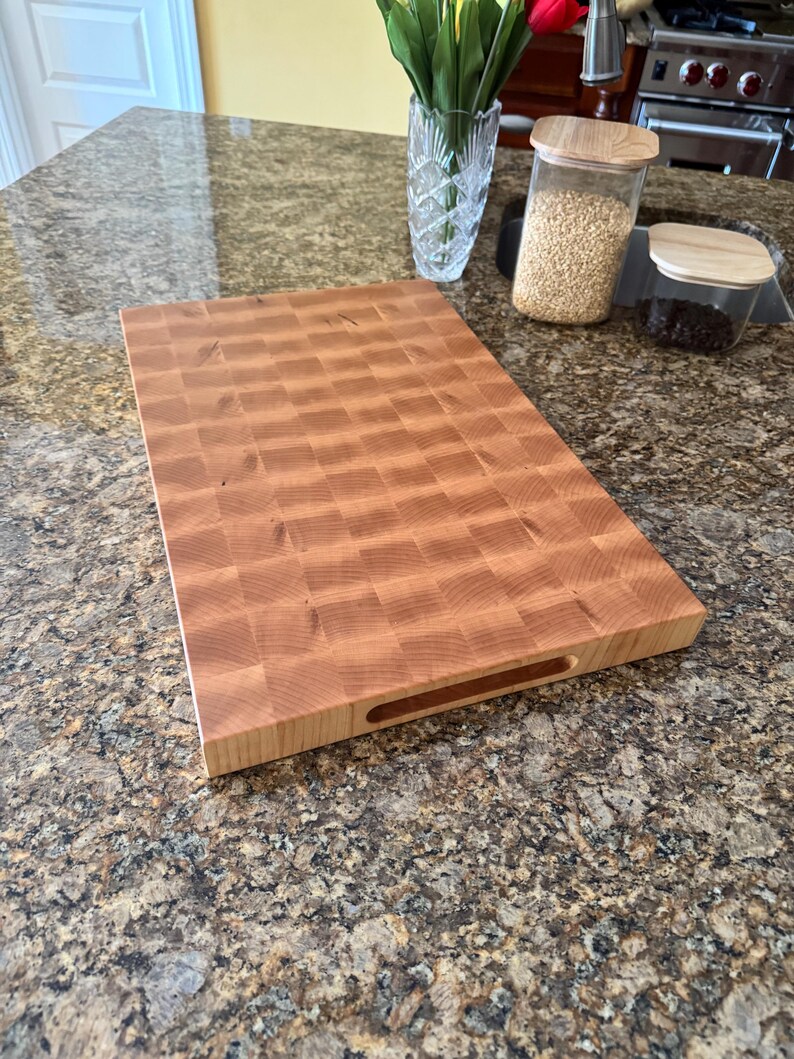 Puede incluir: Una tabla de cortar rectangular de madera de testa con un patr&oacute;n de cuadros de color marr&oacute;n claro. La tabla tiene una superficie lisa y un asa recortada horizontal en el lateral, mostrando la veta natural de la madera en cuadr&iacute;cula.