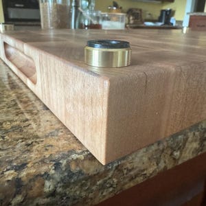 Close-up of end grain hard maple cutting board corner showing routed side handle and non-slip bronze foot with black rubber insert.