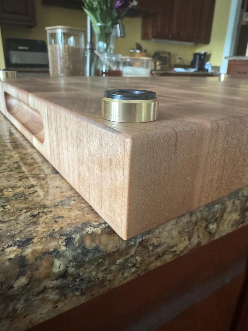 Close-up of end grain hard maple cutting board corner showing routed side handle and non-slip bronze foot with black rubber insert.