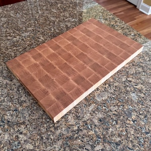 Puede incluir: Una tabla de cortar rectangular de madera de testa en color marr&oacute;n claro. La superficie presenta un patr&oacute;n de cuadr&iacute;cula de peque&ntilde;os bloques de madera cuadrados con texturas de grano visibles, mostrando un aspecto natural y tonal en la parte superior.