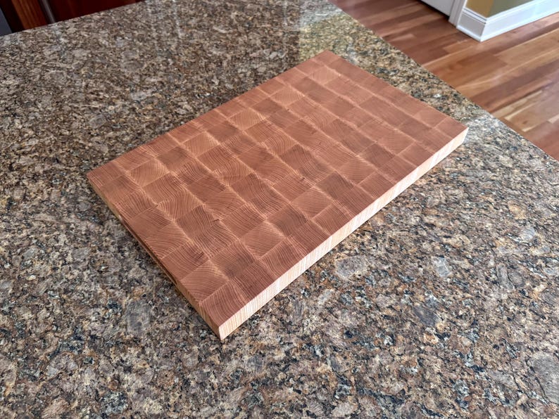 Puede incluir: Una tabla de cortar rectangular de madera de testa en color marr&oacute;n claro. La superficie presenta un patr&oacute;n de cuadr&iacute;cula de peque&ntilde;os bloques de madera cuadrados con texturas de grano visibles, mostrando un aspecto natural y tonal en la parte superior.
