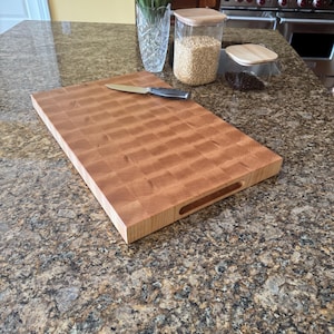 Puede incluir: Una tabla de cortar rectangular de madera de grano final con un patr&oacute;n de cuadros y asas laterales integradas. Un cuchillo de cocina de acero inoxidable con mango negro descansa sobre la superficie de la tabla de madera clara.