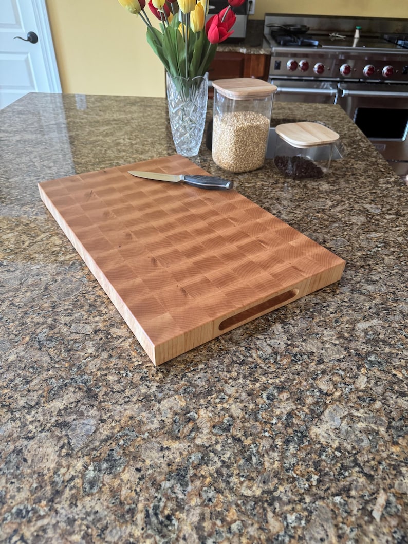 Puede incluir: Una tabla de cortar rectangular de madera de grano final con un patr&oacute;n de cuadros y asas laterales integradas. Un cuchillo de cocina de acero inoxidable con mango negro descansa sobre la superficie de la tabla de madera clara.