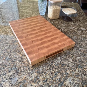Puede incluir: Una tabla de cortar rectangular de madera con construcci&oacute;n de grano final y un patr&oacute;n de cuadr&iacute;cula visible de bloques de madera de color marr&oacute;n claro y medio. La tabla tiene asas laterales integradas en los bordes.