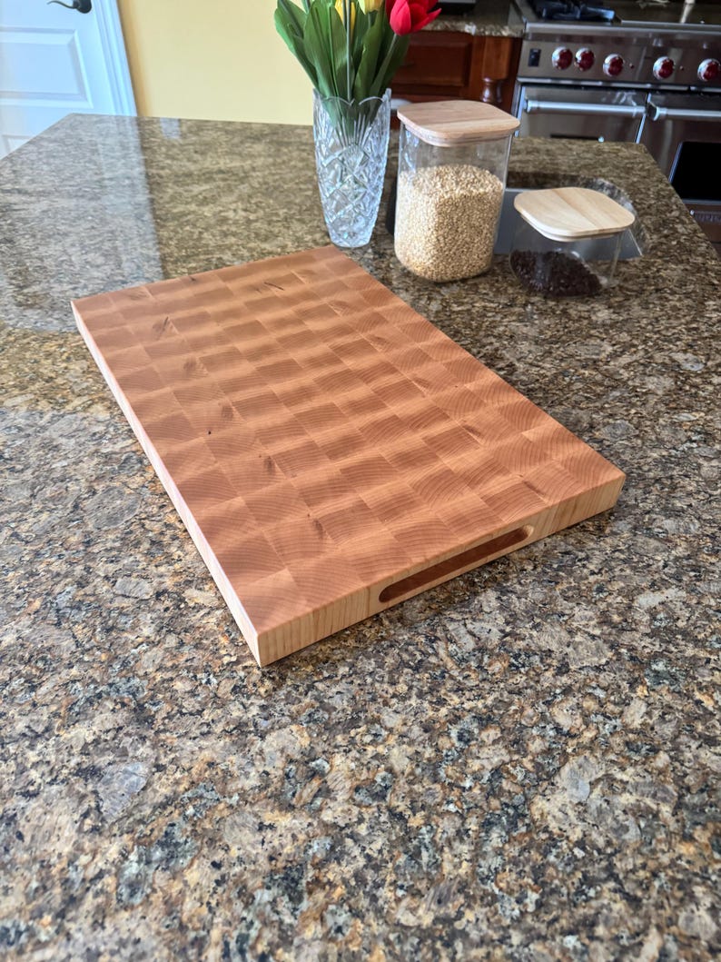 Puede incluir: Una tabla de cortar rectangular de madera con construcci&oacute;n de grano final y un patr&oacute;n de cuadr&iacute;cula visible de bloques de madera de color marr&oacute;n claro y medio. La tabla tiene asas laterales integradas en los bordes.
