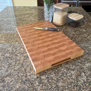 Puede incluir: Una tabla de cortar rectangular de madera de grano final con un patr&oacute;n de cuadros marr&oacute;n claro y un asa empotrada en el lateral. Un cuchillo de cocina de color plateado con mango negro descansa horizontalmente sobre la tabla de madera.
