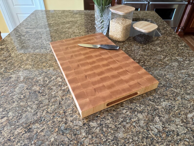 Puede incluir: Una tabla de cortar rectangular de madera de grano final con un patr&oacute;n de cuadros marr&oacute;n claro y un asa empotrada en el lateral. Un cuchillo de cocina de color plateado con mango negro descansa horizontalmente sobre la tabla de madera.
