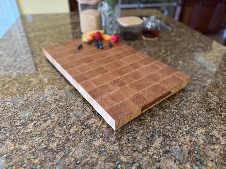 Puede incluir: Una tabla de cortar rectangular de madera de grano final con un patr&oacute;n de cuadr&iacute;cula de bloques de madera de color marr&oacute;n claro y medio. La tabla tiene un perfil grueso con una superficie lisa y una ranura de agarre lateral para facilitar su uso.