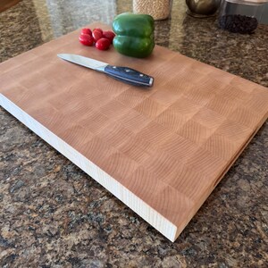 Puede incluir: Una tabla de cortar rectangular de madera de testa con un patr&oacute;n de cuadr&iacute;cula marr&oacute;n claro. Un cuchillo de cocina plateado con mango negro descansa sobre la tabla junto a un pimiento verde grande y varios tomates cherry rojos en grupo.