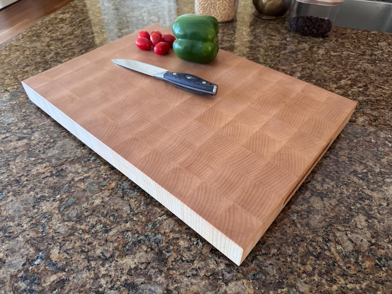 Puede incluir: Una tabla de cortar rectangular de madera de testa con un patr&oacute;n de cuadr&iacute;cula marr&oacute;n claro. Un cuchillo de cocina plateado con mango negro descansa sobre la tabla junto a un pimiento verde grande y varios tomates cherry rojos en grupo.