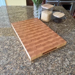 Puede incluir: Una tabla de cortar rectangular de madera de grano final con un patr&oacute;n de cuadros marr&oacute;n claro y un acabado liso. La tabla cuenta con un asa recortada en el lateral, mostrando la textura natural de la veta de la madera y una estructura gruesa.