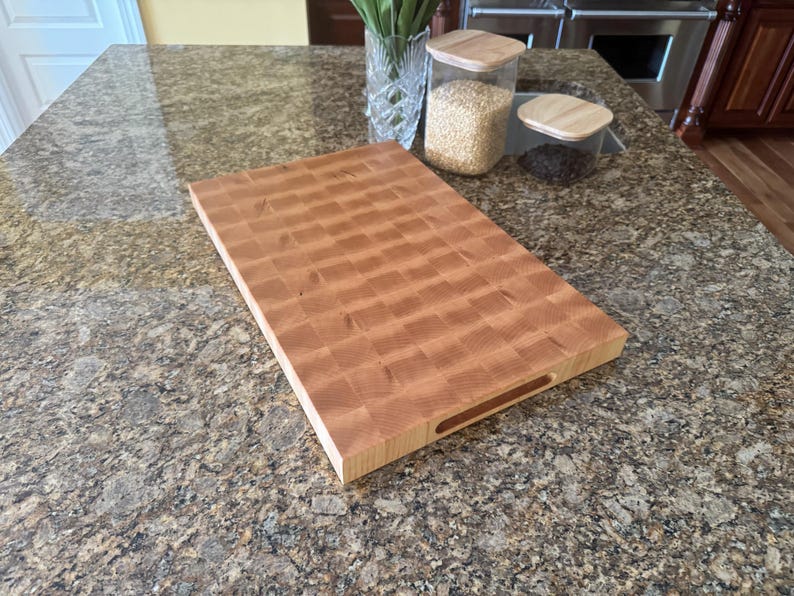 Puede incluir: Una tabla de cortar rectangular de madera de grano final con un patr&oacute;n de cuadros marr&oacute;n claro y un acabado liso. La tabla cuenta con un asa recortada en el lateral, mostrando la textura natural de la veta de la madera y una estructura gruesa.