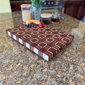 May include: A rectangular end-grain cutting board featuring a geometric pattern of dark brown, medium brown, and light tan wood blocks. The board has a thick construction with visible layered wood strips along the side edges and a smooth surface.