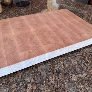 Puede incluir: Una tabla de cortar rectangular de madera clara con veta de testa. La superficie presenta un patr&oacute;n de cuadr&iacute;cula de peque&ntilde;os bloques de madera cuadrados, mientras que el borde lateral muestra la veta vertical de las piezas apiladas.