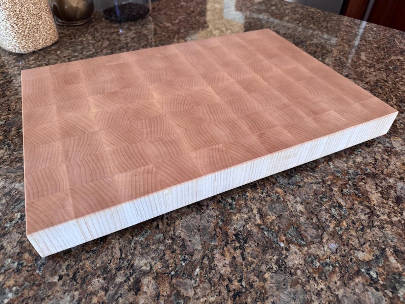 Puede incluir: Una tabla de cortar rectangular de madera clara con veta de testa. La superficie presenta un patr&oacute;n de cuadr&iacute;cula de peque&ntilde;os bloques de madera cuadrados, mientras que el borde lateral muestra la veta vertical de las piezas apiladas.