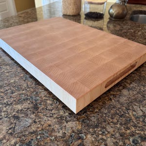 Puede incluir: Una tabla de cortar rectangular de madera de testa en color beige claro con un patr&oacute;n de cuadr&iacute;cula visible de bloques de madera. La tabla presenta un perfil grueso y un asa empotrada tallada en el lateral para facilitar su agarre.