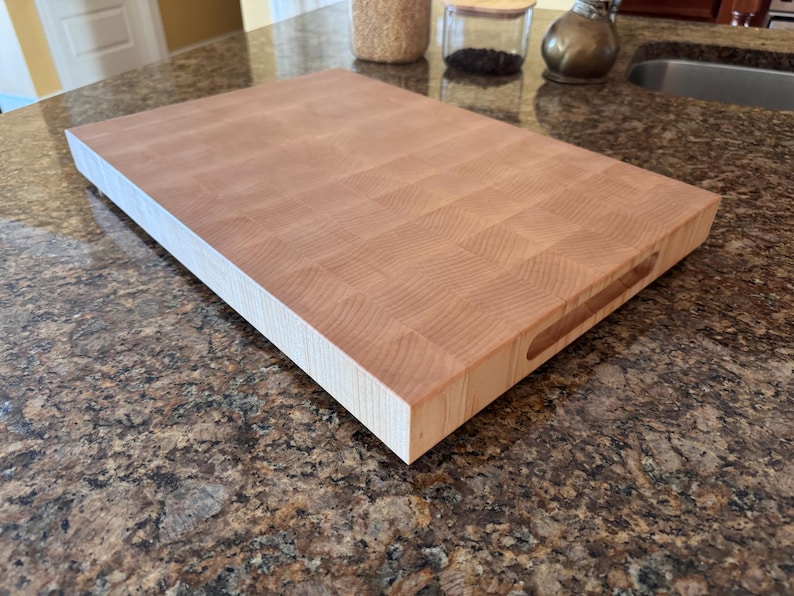 Puede incluir: Una tabla de cortar rectangular de madera de testa en color beige claro con un patr&oacute;n de cuadr&iacute;cula visible de bloques de madera. La tabla presenta un perfil grueso y un asa empotrada tallada en el lateral para facilitar su agarre.