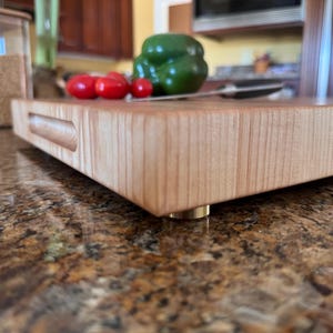 Puede incluir: Una tabla de cortar rectangular de madera clara con un asa empotrada en el lateral y peque&ntilde;os pies met&aacute;licos. La tabla presenta una veta suave y est&aacute; colocada horizontalmente, mostrando su grosor y su construcci&oacute;n s&oacute;lida.