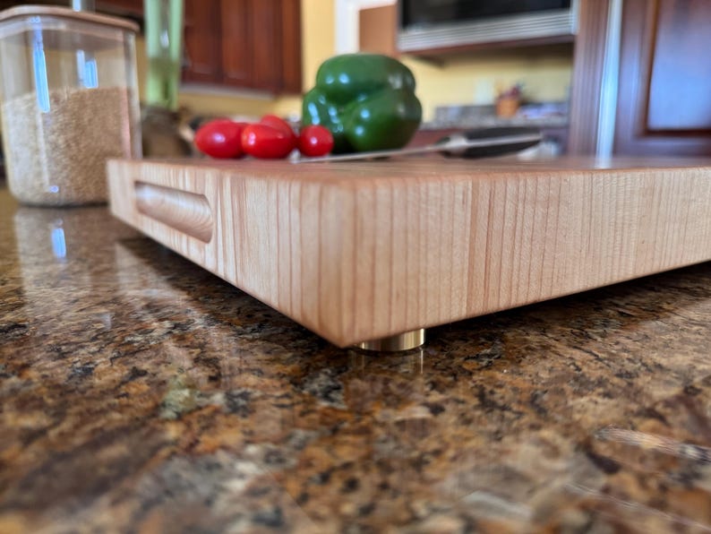 Puede incluir: Una tabla de cortar rectangular de madera clara con un asa empotrada en el lateral y peque&ntilde;os pies met&aacute;licos. La tabla presenta una veta suave y est&aacute; colocada horizontalmente, mostrando su grosor y su construcci&oacute;n s&oacute;lida.