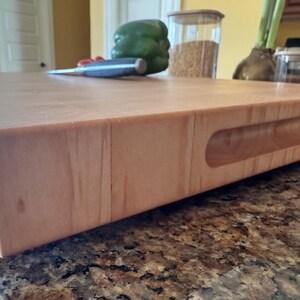 Puede incluir: Una tabla de cortar rectangular y gruesa de madera clara con un acabado liso y lijado. La tabla presenta un asa horizontal empotrada tallada en el lateral, mostrando un patr&oacute;n de veta de madera natural en su construcci&oacute;n de bloque s&oacute;lido.