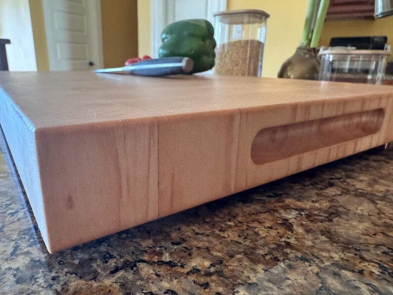Puede incluir: Una tabla de cortar rectangular y gruesa de madera clara con un acabado liso y lijado. La tabla presenta un asa horizontal empotrada tallada en el lateral, mostrando un patr&oacute;n de veta de madera natural en su construcci&oacute;n de bloque s&oacute;lido.