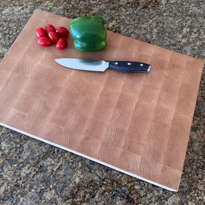 Puede incluir: Una tabla de cortar rectangular de madera de testa con un patr&oacute;n de cuadr&iacute;cula. Un cuchillo de cocina plateado con mango negro descansa sobre la tabla junto a un pimiento verde entero y cinco tomates cherry rojos brillantes.