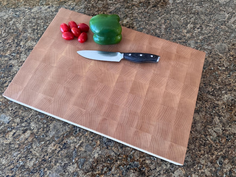 Puede incluir: Una tabla de cortar rectangular de madera de testa con un patr&oacute;n de cuadr&iacute;cula. Un cuchillo de cocina plateado con mango negro descansa sobre la tabla junto a un pimiento verde entero y cinco tomates cherry rojos brillantes.