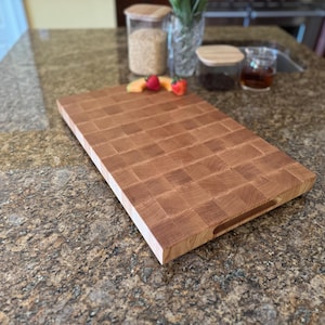 Puede incluir: Una tabla de cortar rectangular de madera de grano final con un patr&oacute;n de cuadr&iacute;cula de bloques de madera de color marr&oacute;n claro y medio. La tabla presenta una superficie lisa y plana y una muesca de agarre en el lateral, mostrando la veta natural.