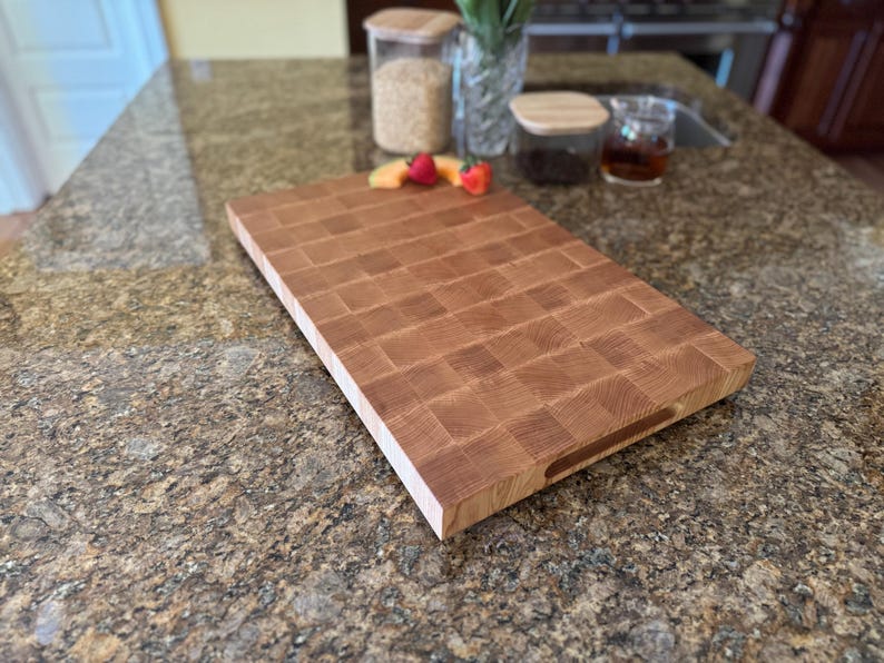 Puede incluir: Una tabla de cortar rectangular de madera de grano final con un patr&oacute;n de cuadr&iacute;cula de bloques de madera de color marr&oacute;n claro y medio. La tabla presenta una superficie lisa y plana y una muesca de agarre en el lateral, mostrando la veta natural.