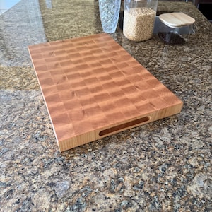 Puede incluir: Una tabla de cortar rectangular de madera de grano final con un patr&oacute;n de cuadros marr&oacute;n claro y un acabado liso. La tabla presenta un asa empotrada en el lateral, mostrando la veta natural de la madera y una construcci&oacute;n gruesa y resistente.