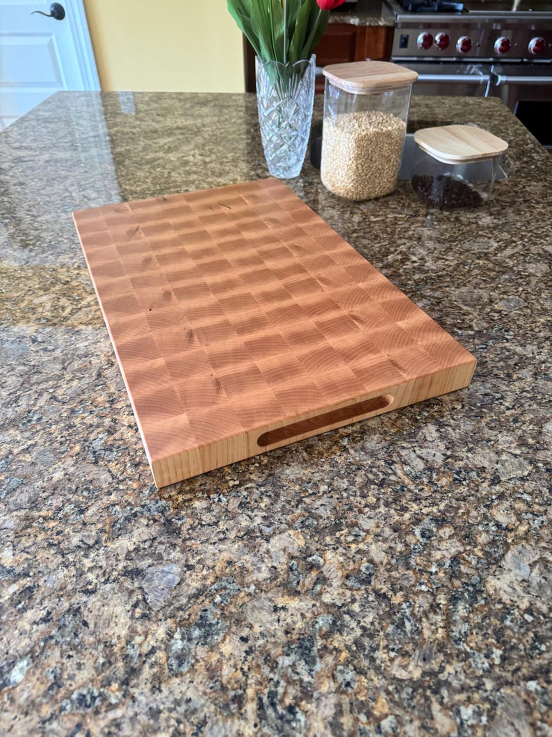 Puede incluir: Una tabla de cortar rectangular de madera de grano final con un patr&oacute;n de cuadros marr&oacute;n claro y un acabado liso. La tabla presenta un asa empotrada en el lateral, mostrando la veta natural de la madera y una construcci&oacute;n gruesa y resistente.