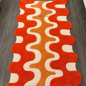 May include: A rectangular rug with a wavy, scalloped edge featuring a repeating pattern of vertical, undulating stripes in vibrant orange, burnt orange, and cream. The rug has a tufted texture and includes the text BIKHA ENTERPRISES in the centre.