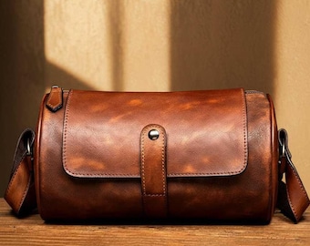 May include: A brown leather barrel bag with a flap closure and a shoulder strap. The bag is made of distressed leather and has a silver-colored button closure. The bag is sitting on a wooden surface.