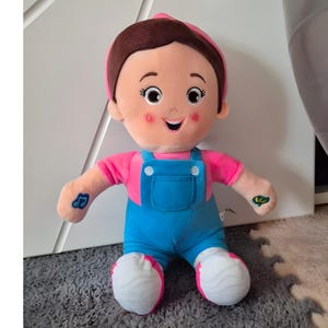 Può includere: Una bambola di peluche sorridente, con una maglietta rosa, una salopette blu e un berretto rosa. La bambola ha i capelli castani e scarpe bianche con dettagli rosa. Disegni di note musicali e foglie sulle braccia.