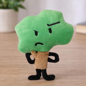Pode incluir: Um brinquedo de pelúcia em forma de florete de brócolis verde com olhos pretos e uma carranca. Possui um corpo marrom e braços e pernas pretas, dando-lhe uma aparência de desenho animado. O brinquedo foi projetado para ficar em pé.