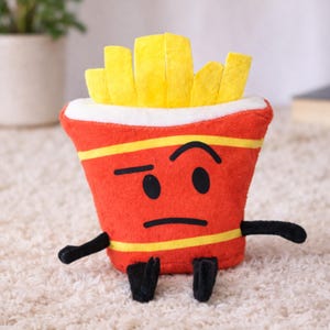 Pode incluir: Um brinquedo de pelúcia em forma de um recipiente de batatas fritas. O recipiente vermelho tem listras amarelas e um rosto com uma expressão cética. As batatas fritas são amarelas e saem do topo. Braços e pés pretos completam o design.