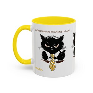 Puede incluir: Taza de café de cerámica blanca con interior y asa amarillos. La taza presenta una ilustración de dibujos animados de un gato negro gruñón con una corbata a rayas amarillas y blancas. El texto en la taza dice: "Coffee: Because adulting is hard."
