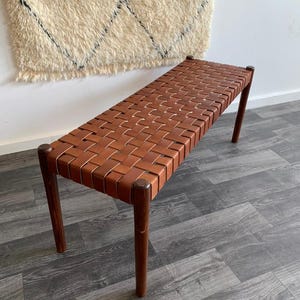 May include: A brown leather bench with a woven seat and four wooden legs. The bench is approximately 48 inches long and is set against a white wall with a cream-colored rug.