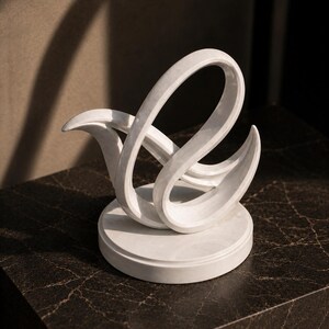May include: A white marble sculpture featuring a smooth, curved, abstract form that resembles a stylized bird or swan. The piece is mounted on a circular base, showcasing a polished finish with subtle grey veining throughout the stone structure.