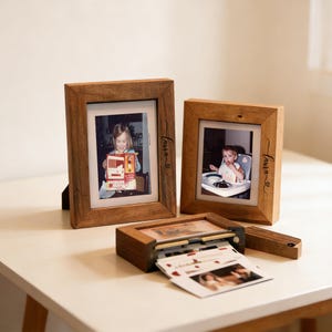 May include: Three wooden picture frames with photos and a small photo printer. The frames are rectangular with a natural wood finish. The photos show a young child. The name "Laura" is written on the frames.