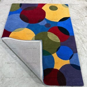 May include: A rectangular rug with an abstract design of overlapping circles in various colours. The rug's palette includes blue, yellow, red, green, and purple. The reverse side of the rug is visible, showing a grey backing.