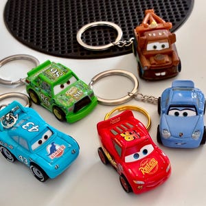 Può includere: Un set di cinque portachiavi raffiguranti auto in miniatura tratte dal film Cars. Il set include un carro attrezzi marrone, un'auto da corsa verde con il numero 86, un'auto da corsa azzurra con il numero 43, un'auto rossa con il numero 95 e un'auto sportiva azzurra.