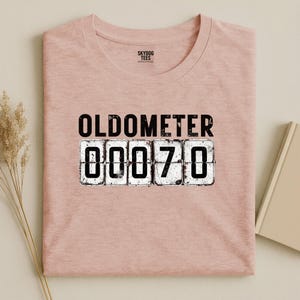 May include: A light pink t-shirt with the word "OLDOMETER" above the number "00070" in a distressed font. The shirt has a crew neck and the brand "SKYDOG TEES" is printed on the tag.
