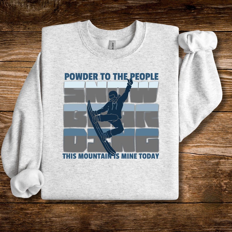 May include: A light gray sweatshirt with a snowboarder graphic in navy blue and light blue. The text "POWDER TO THE PEOPLE" and "SNOW BOARDING" are above the snowboarder, and "THIS MOUNTAIN IS MINE TODAY" is below.
