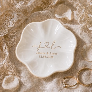 May include: A white ceramic ring dish with a scalloped, flower-like edge. The center features gold-colored text that reads j heart l, Jessica & Lucas, and the date 12.06.2026. The dish has a smooth, glossy finish and a shallow, rounded basin.